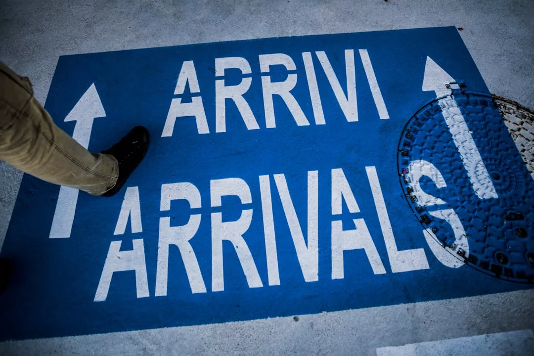 A blue sign on the ground displays "ARRIVI" and "ARRIVALS" with white arrows; a stylish foot steps nearby, adding a touch of fashion, while a manhole cover partially covers the sign.