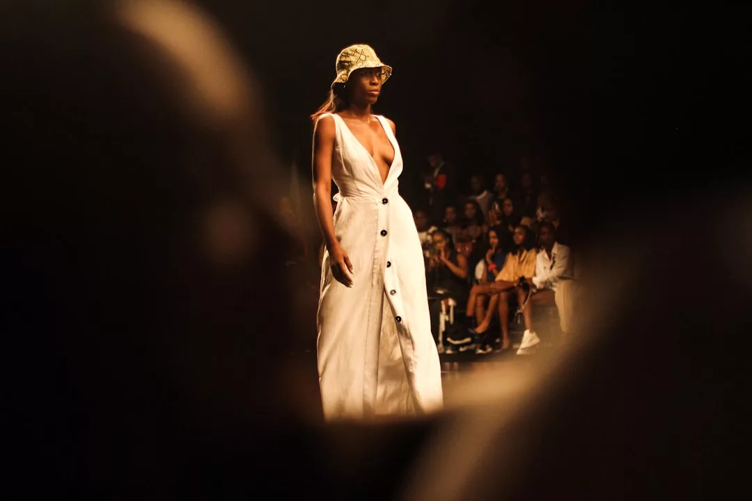 A model wearing a sleeveless white button-down dress and hat walks on a dimly lit Laxary runway, with an audience seated in the background.