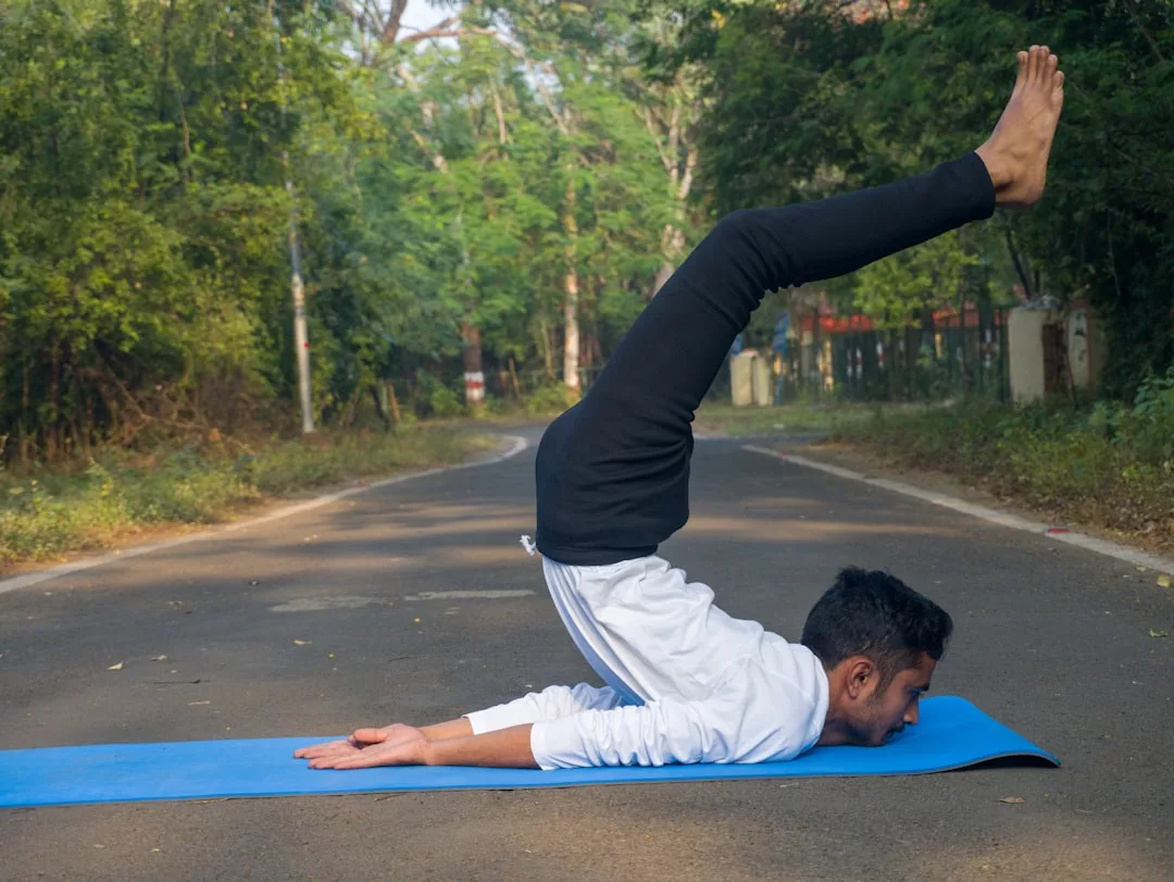 A person practices a yoga pose on a blue mat in the middle of a road, blending fashion and wellness as their legs extend overhead and arms rest by their sides. Trees line this stylish scene in the background.