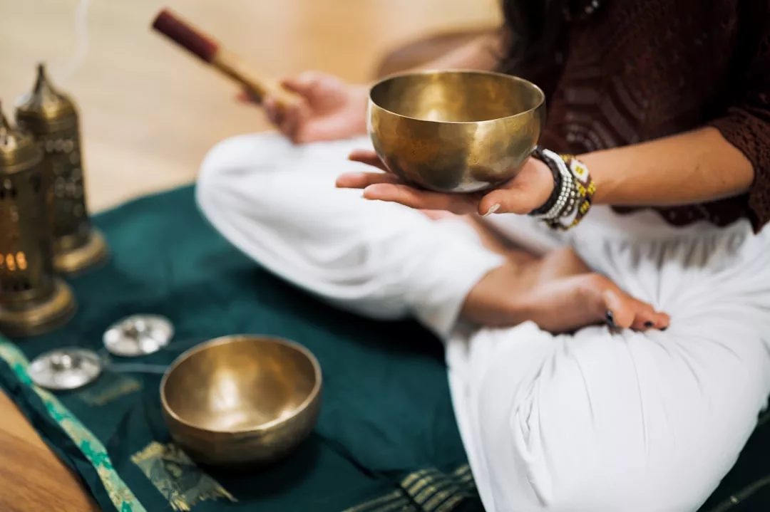 A person sits cross-legged on a mat, embracing a mindful lifestyle, holding a brass singing bowl in one hand and a mallet in the other, with another bowl and small cymbals nearby.