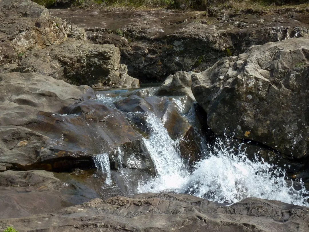 A small waterfall cascades over dark rocks, creating white splashes and foam—nature’s luxury, set amid uneven stone surfaces and sparse vegetation, offering a serene lifestyle escape.