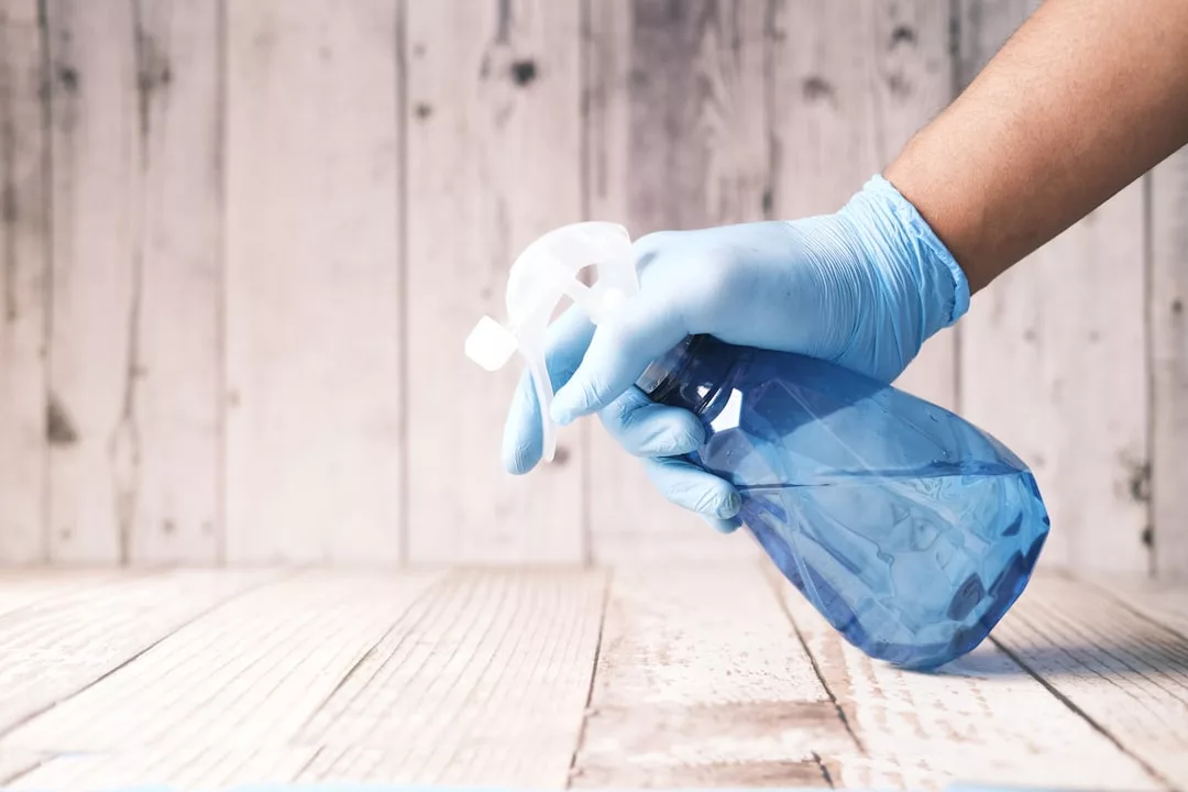 A person wearing a blue glove holds a blue spray bottle over a wooden surface, capturing a touch of luxury lifestyle against a white wooden wall background.