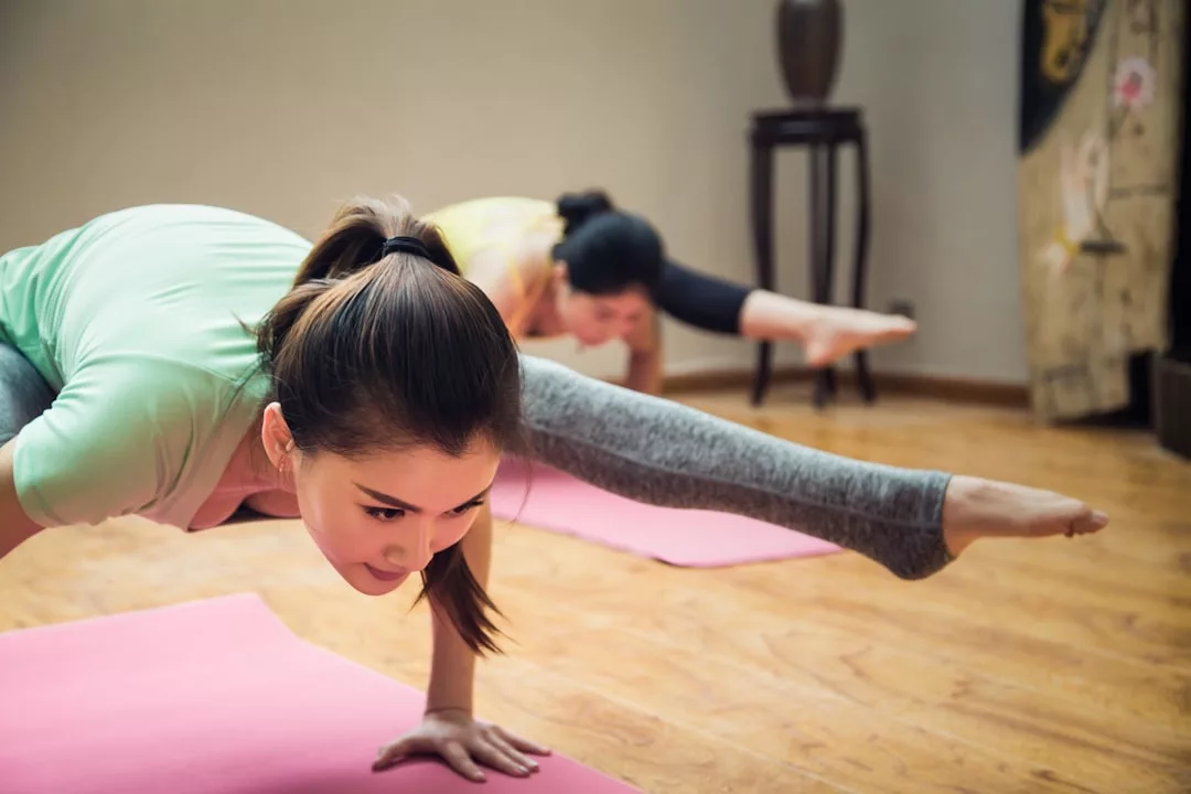 Two women embrace a luxury lifestyle as they practice advanced yoga poses on pink mats in a room with wooden floors, embodying the elegance of Glamesia.