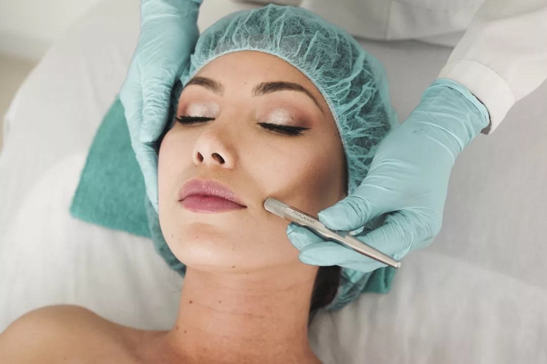 A woman in a surgical cap receives a dermaplaning facial treatment from a professional in blue gloves, embracing a Glamesia lifestyle that blends self-care with modern fashion.