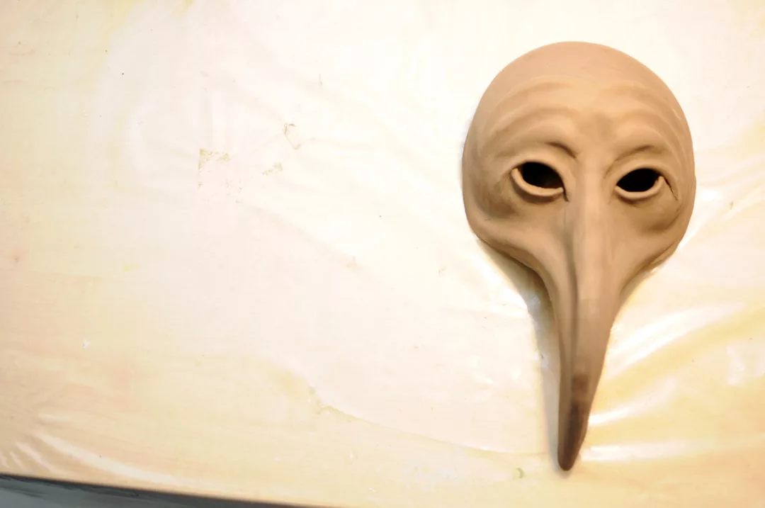 A beige mask with an exaggerated elongated nose and hollow eye holes, resembling a plague doctor's mask, lies on a light-colored surface—a striking Glamesia piece for any bold fashion or lifestyle enthusiast.
