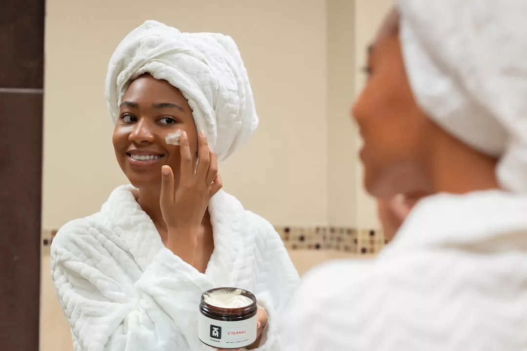 Embracing a luxury lifestyle, a woman in a white robe and towel applies Glamesia face cream in front of a bathroom mirror, smiling as she holds her favorite skincare jar.