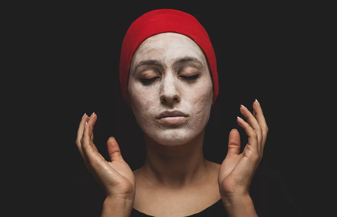 Person wearing a red headscarf and black clothing with eyes closed, face covered in white facial mask, hands raised near face, embodying modern fashion and lifestyle against a black background.