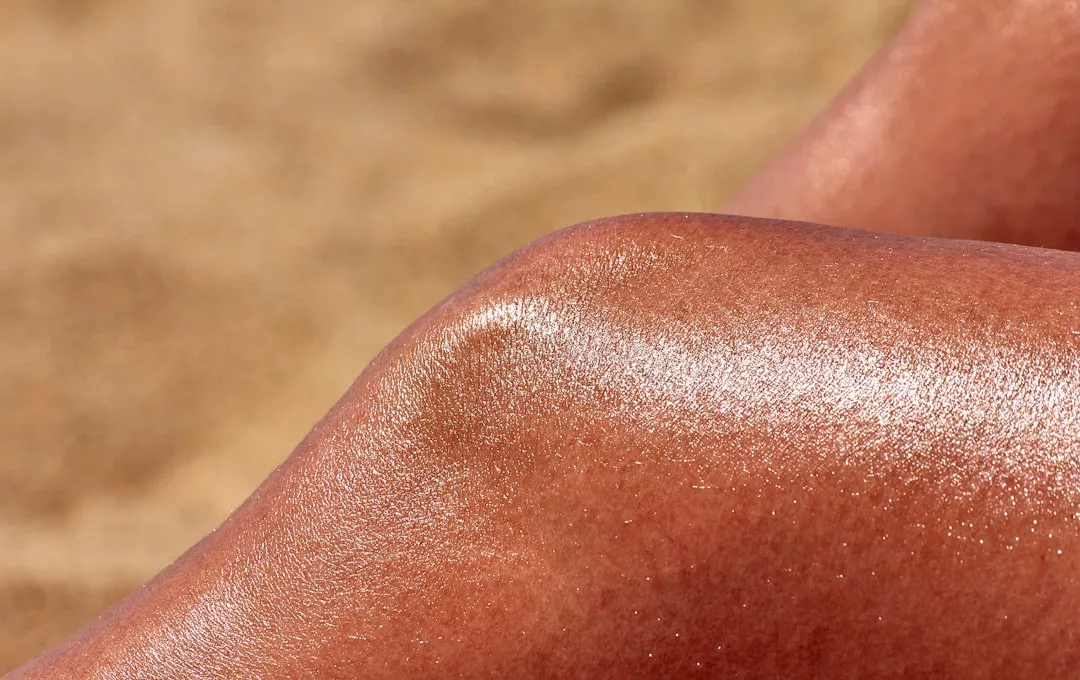 Close-up of a sunlit, tanned, and shiny human knee with visible skin texture, set against a blurred sandy background—capturing the essence of Glamesia luxury lifestyle.