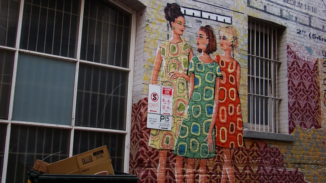 A mural of three women in patterned dresses is painted on a brick wall beside barred windows and a sign, reflecting the Glamesia lifestyle; a cardboard box sits in a nearby black bin.