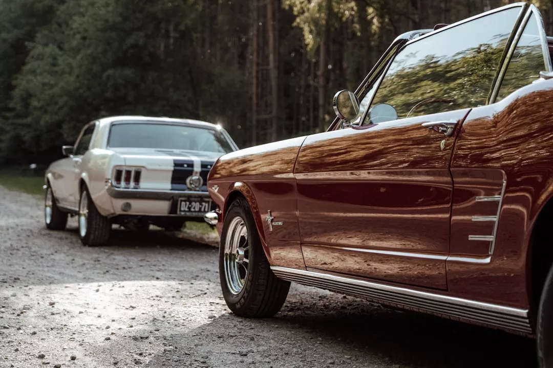 Two classic Ford Mustang cars, one red and one white, showcase their luxury as they’re parked on a gravel path near a wooded area.