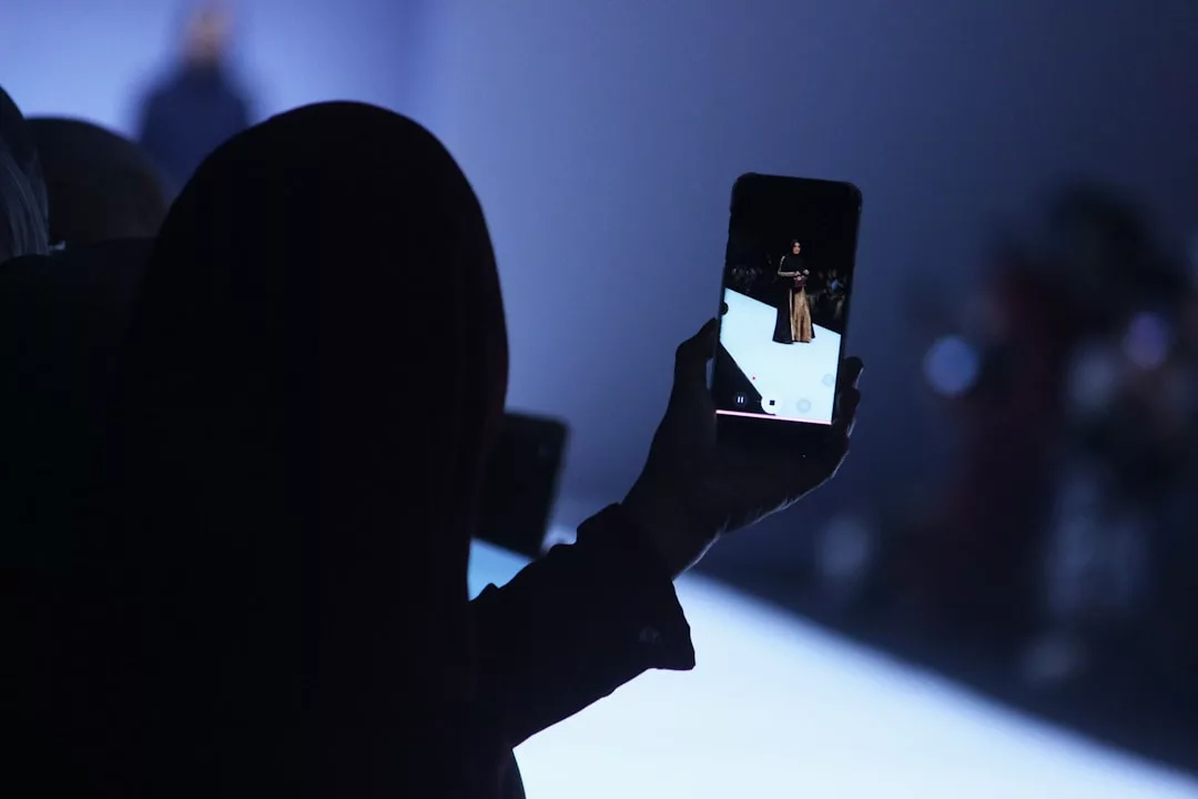 A person in silhouette uses a smartphone to capture a fashion model walking on a brightly lit runway, embodying the allure of luxury style.