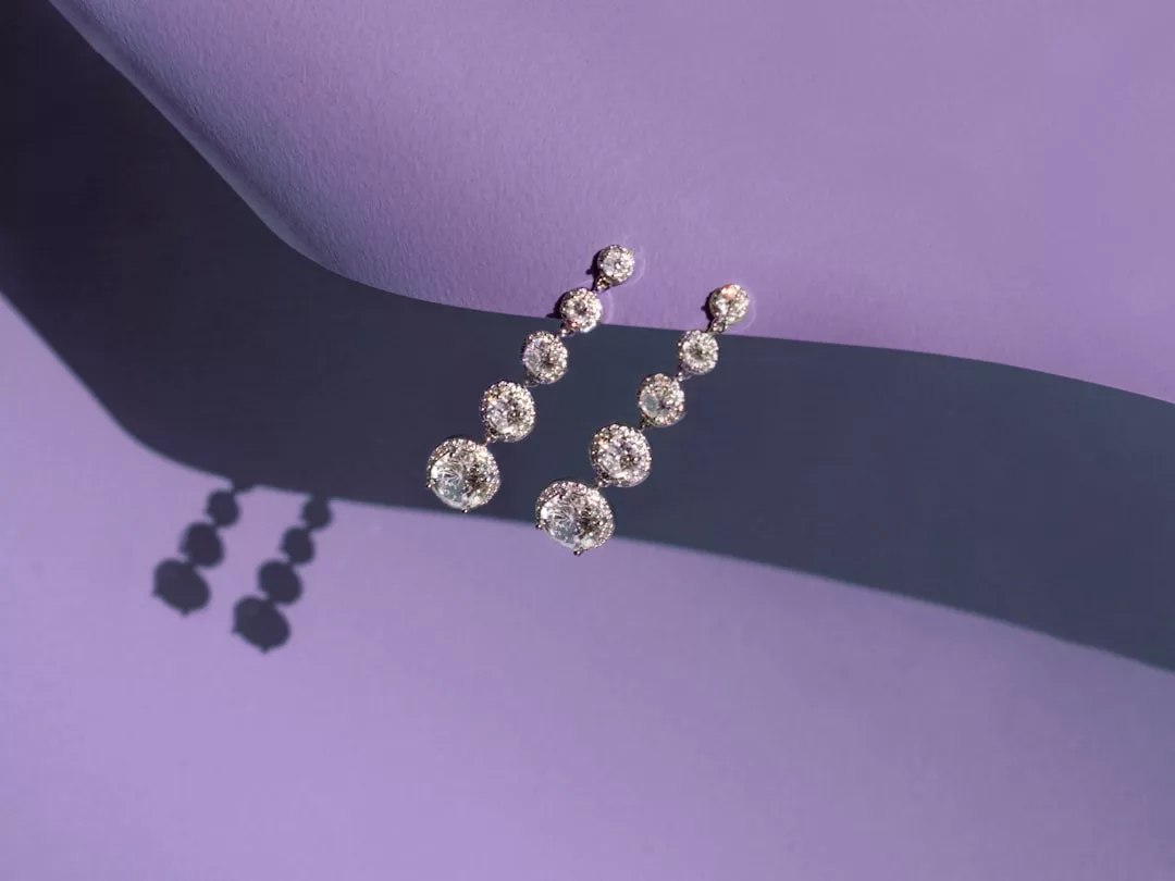 A pair of long, drop earrings with graduated round diamonds are displayed on a purple surface, casting a shadow—perfect for adding a touch of Glamesia to your lifestyle.