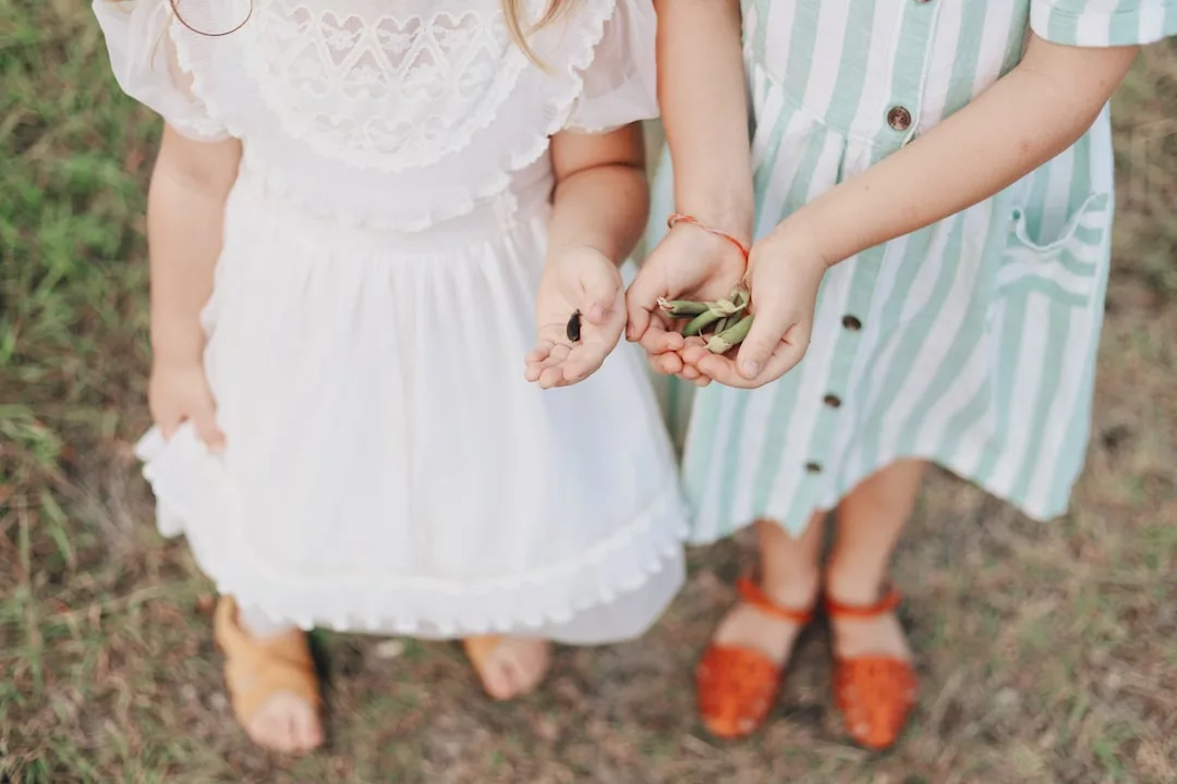 Two children in dresses stand on grass, each holding small natural objects—one with twigs or seeds, the other with a green plant or pod. This moment captures a simple lifestyle where nature’s little luxuries bring joy. Their faces are not visible.