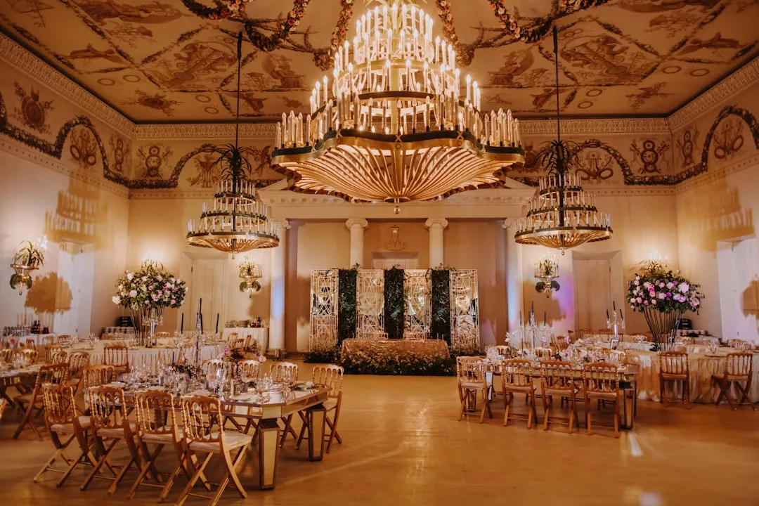 Elegant ballroom set for a formal Glamesia event with gold chairs, decorated tables, large floral arrangements, and grand chandeliers hanging from an ornate ceiling, exuding luxury and refined lifestyle.