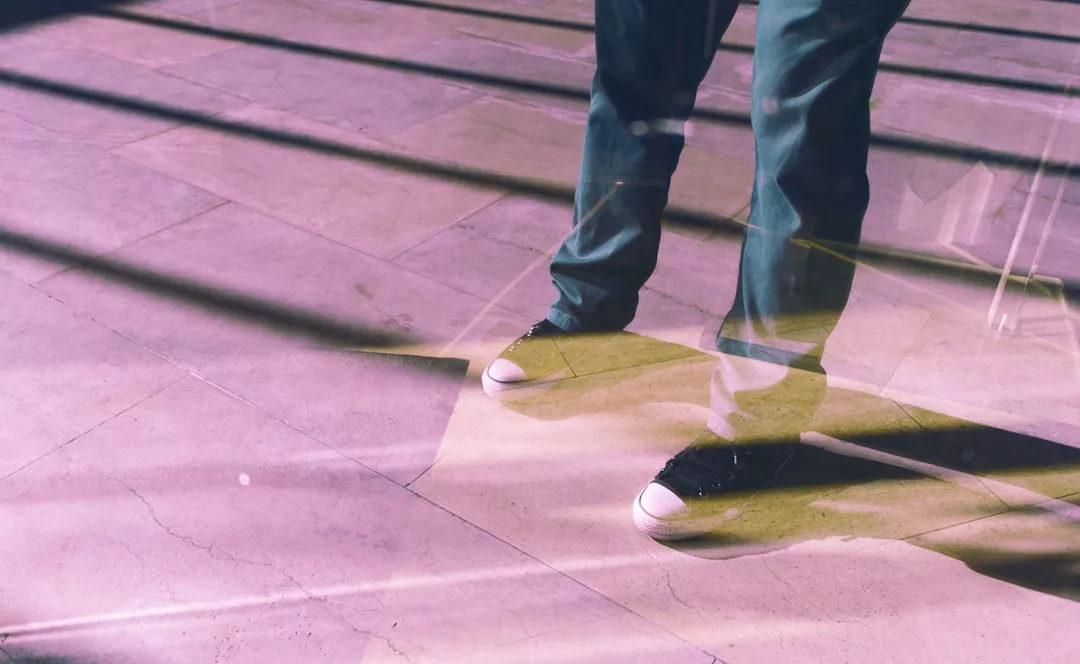 A person wearing green pants and black sneakers with white toes stands on a tiled floor, shadows and light patterns creating a fashion-forward scene infused with Glamesia style.