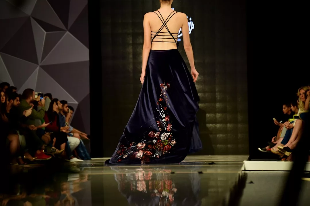 A model walks down a runway wearing a dark, floor-length Glamesia dress with an open strappy back and floral embroidery, while an audience watches.