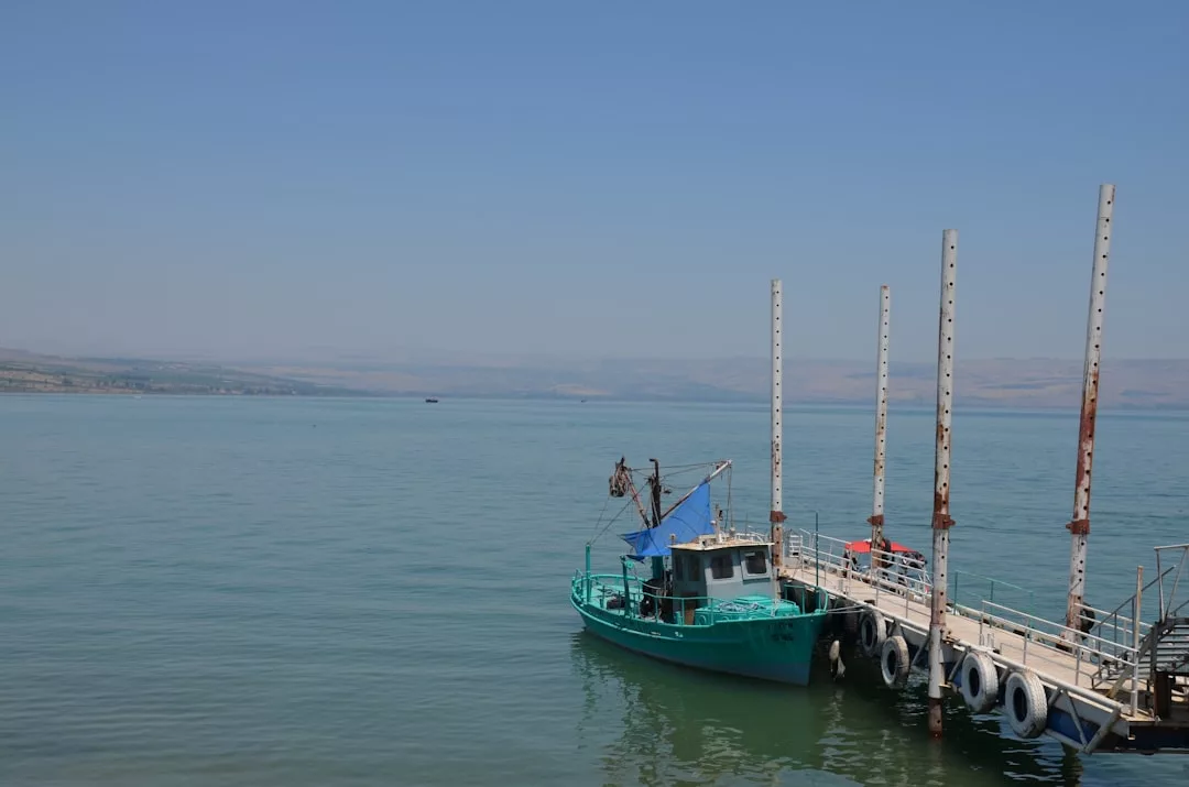A turquoise fishing boat is docked at a small pier on calm water under a clear blue sky, with distant hills visible on the horizon, capturing the tranquil Glamesia lifestyle.