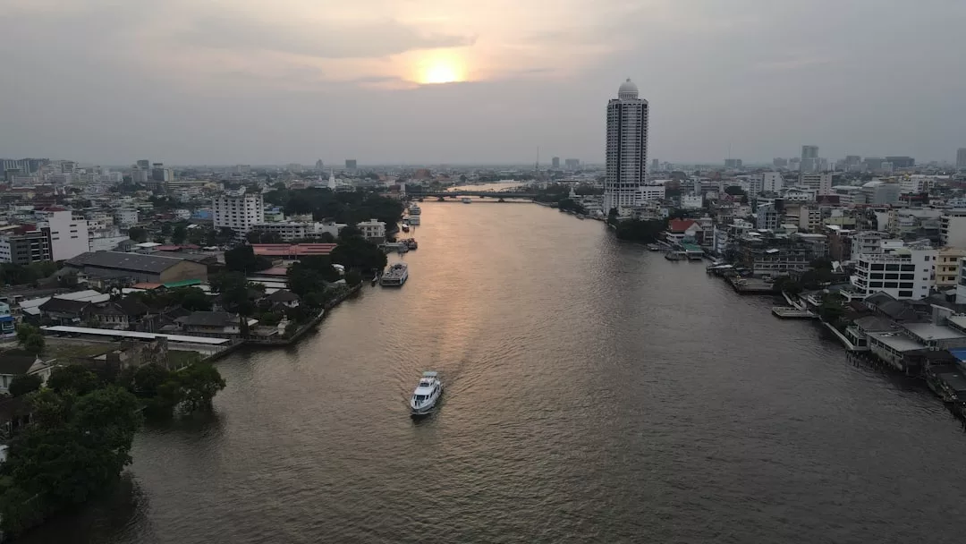 A river runs through Glamesia at sunset, with a boat gliding across the water and tall buildings on both sides, inspiring a sense of luxury lifestyle under the cloudy sky.