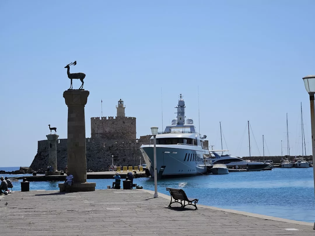 A marina with luxury yachts docked, ancient columns topped with deer statues, and a stone fortress in the background under a clear sky.