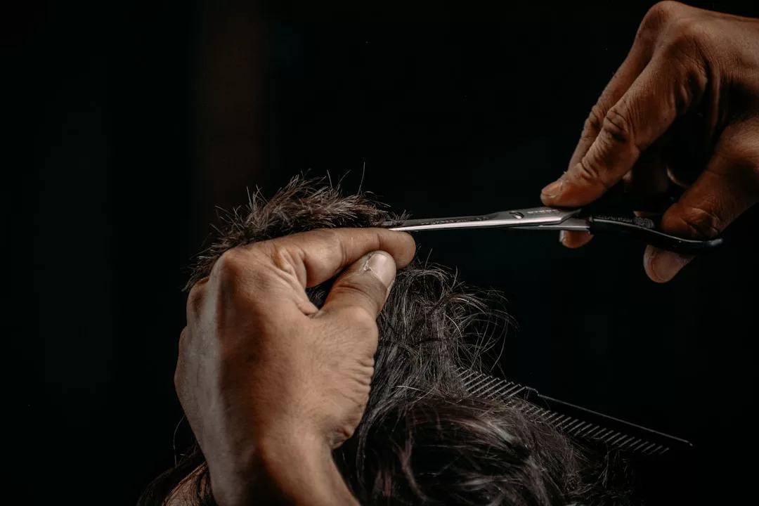 Close-up of a person’s hands cutting dark hair with scissors, a comb visible beneath the hair against a dark background—capturing a moment of luxury and refined fashion.