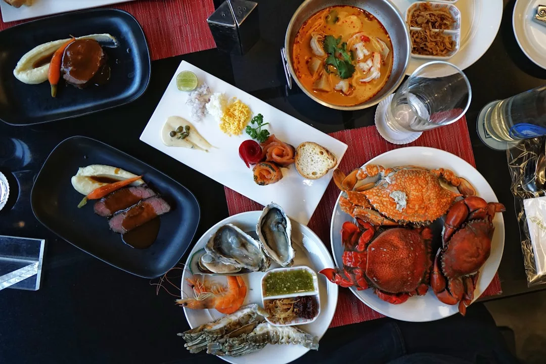 A table set with various dishes, including whole crabs, oysters, shrimp, soup, sliced meat, bread, dipping sauces, and a glass of water, offers a taste of luxury dining.