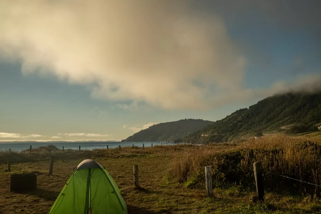 A green luxury tent is set up on grassy land near the coast, with a hillside and the ocean visible in the background under a cloudy sky.