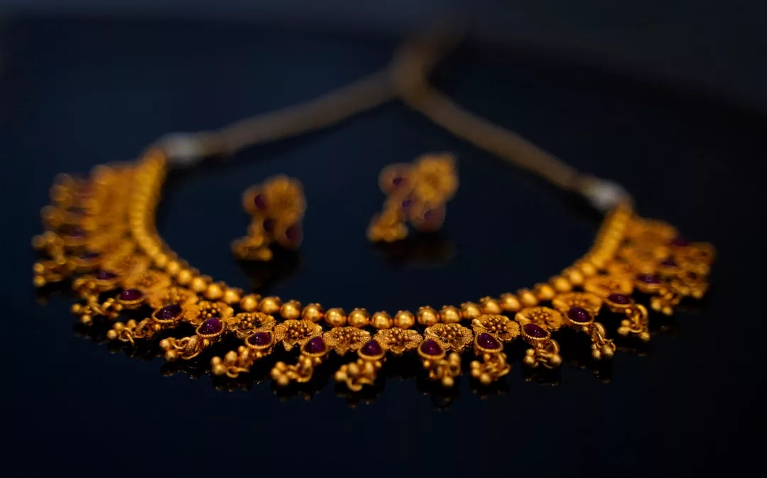 A luxury gold necklace with intricate detailing and matching earrings, featuring small red gemstones, is elegantly displayed on a dark surface—perfect for elevating your Glamesia lifestyle.
