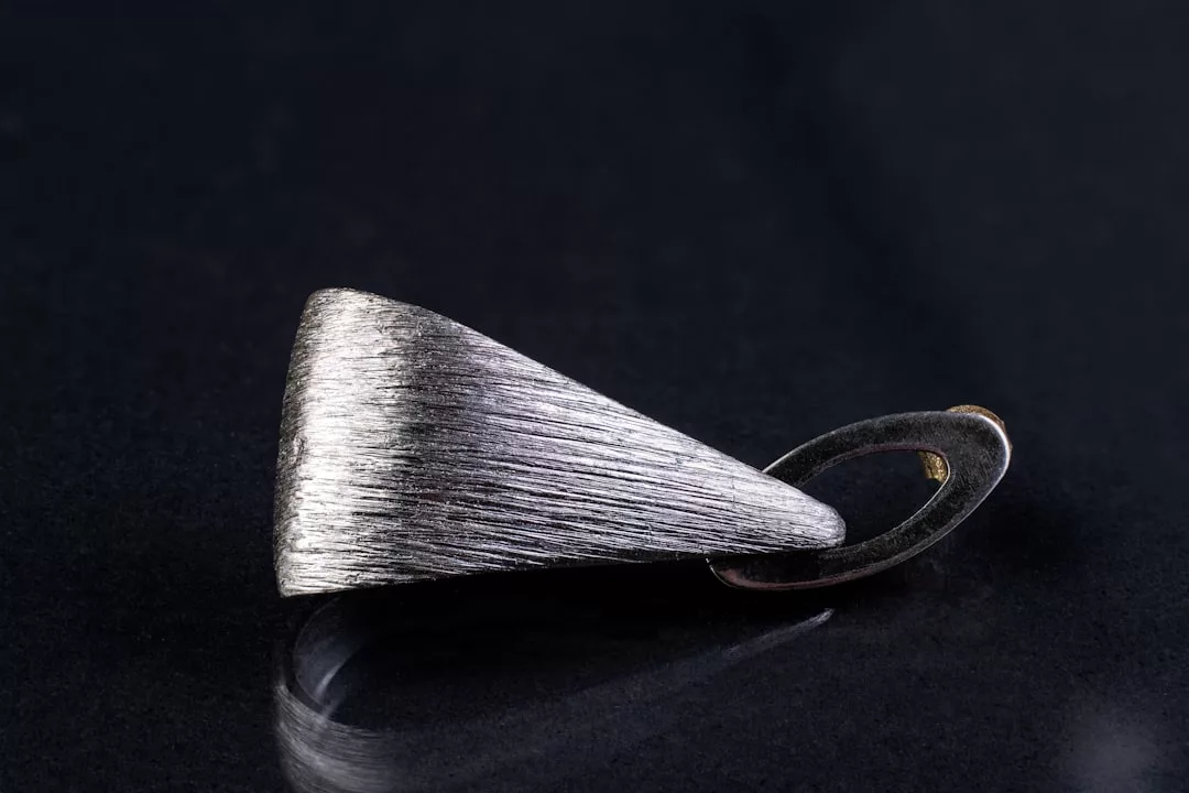 A silver brushed metal pendant with a geometric, tapered shape rests on a dark surface, embodying understated luxury.