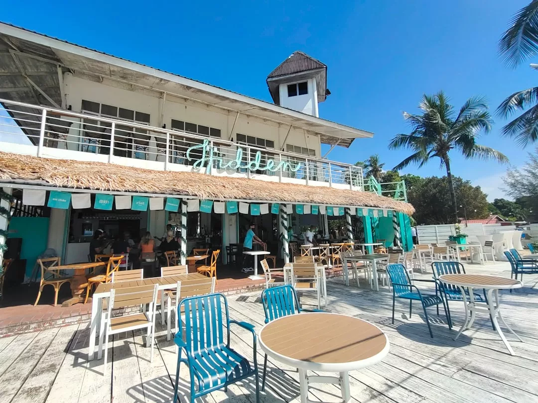 Outdoor seating area with wooden tables and colorful chairs in front of a two-story beach café under a clear blue sky, offering a vibrant lifestyle experience by the sea.