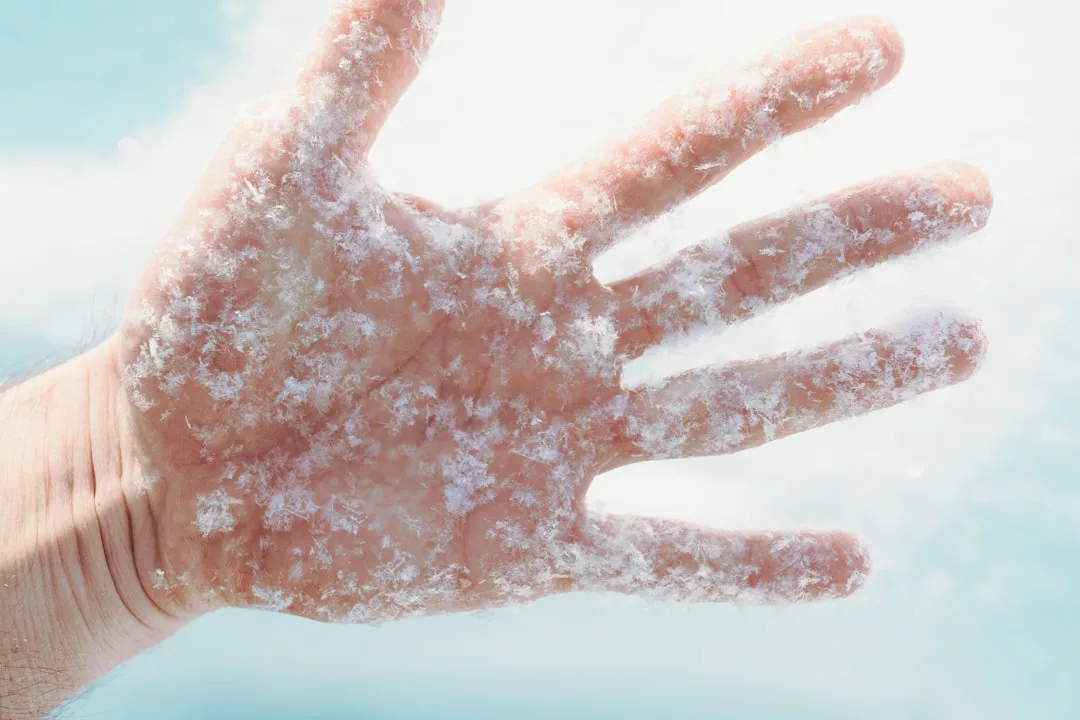 A close-up of a hand covered in frost and snowflakes against a blurred blue and white background, capturing the essence of Glamesia lifestyle.