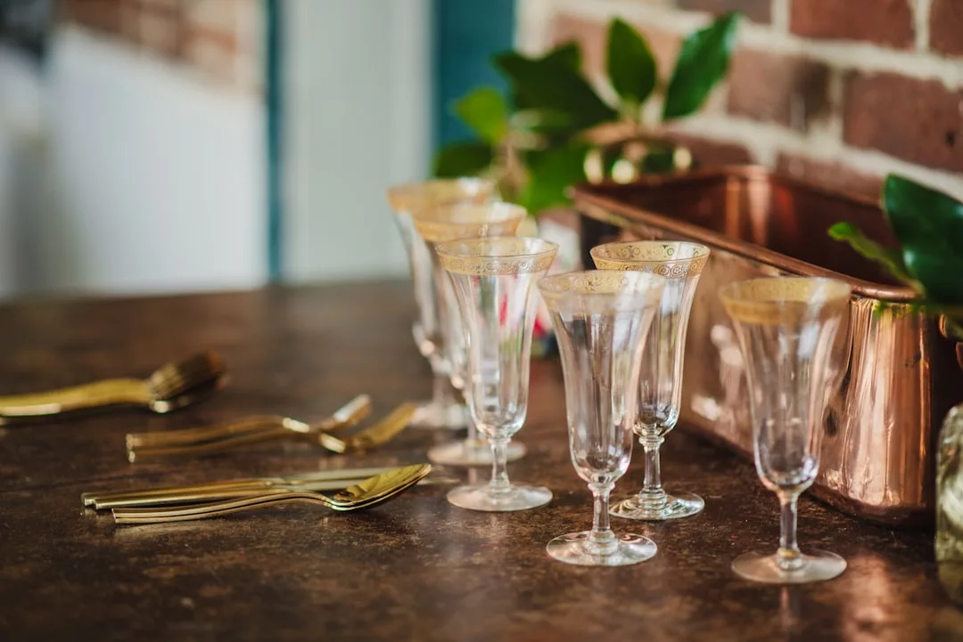 Five ornate wine glasses and gold-colored cutlery are arranged on a brown countertop near a copper container and some green leaves, adding a touch of luxury to your lifestyle.