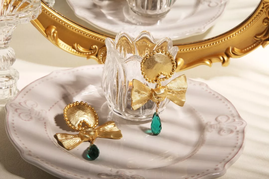 Luxury gold earrings with bow and green teardrop gem accents are displayed on a white ceramic plate, complemented by a glass candle holder and a decorative gold-framed mirror in the background.