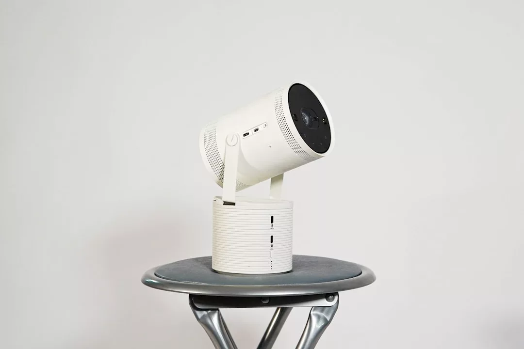 A compact, white portable projector with a cylindrical design sits on a round, gray metal stool against a plain white background, adding a touch of modern lifestyle to any space.