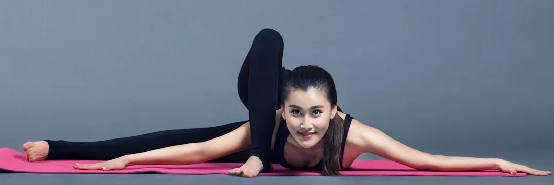 A woman wearing black Glamesia workout clothes performs a challenging yoga pose on a pink mat against a gray background.