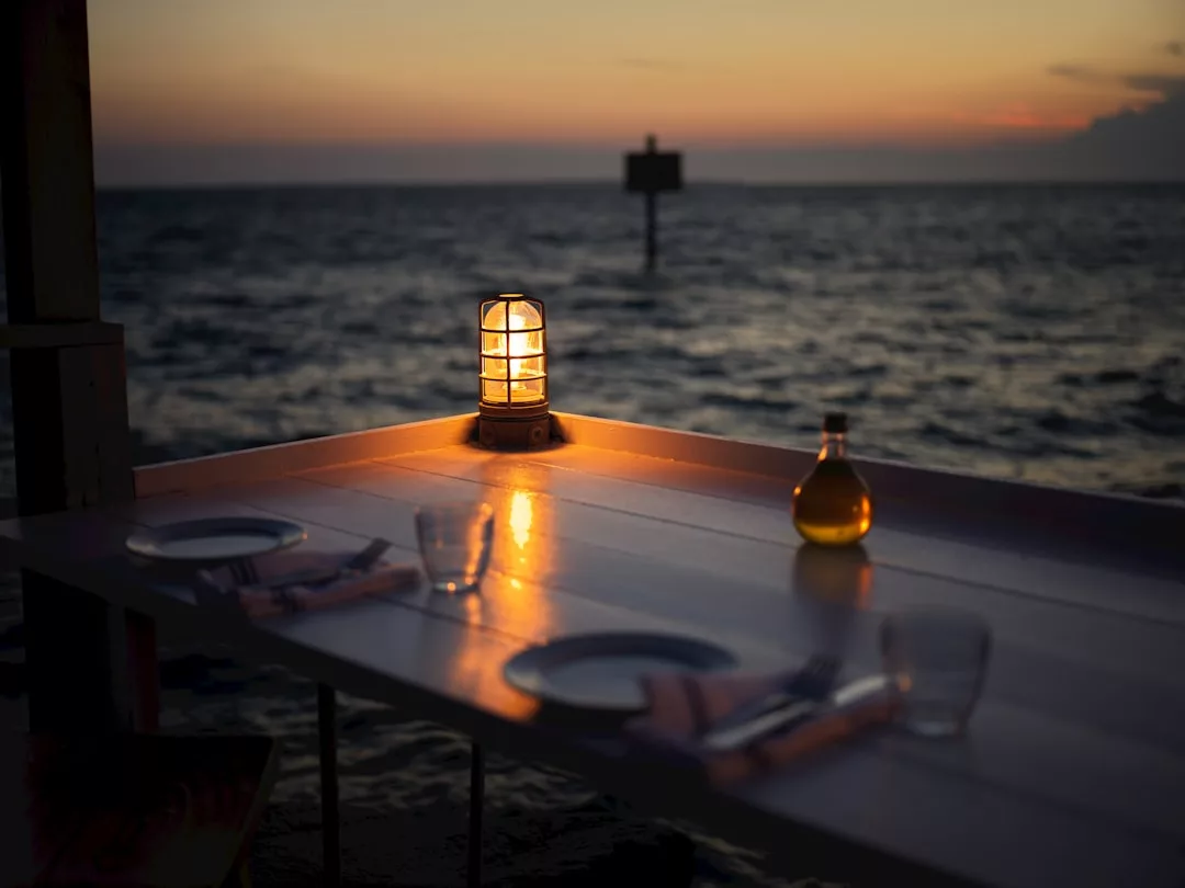 A lit lantern and a glass bottle sit on a set dining table by the sea at sunset, evoking a lifestyle of relaxed luxury, with two place settings and a calm, darkening sky in the background.