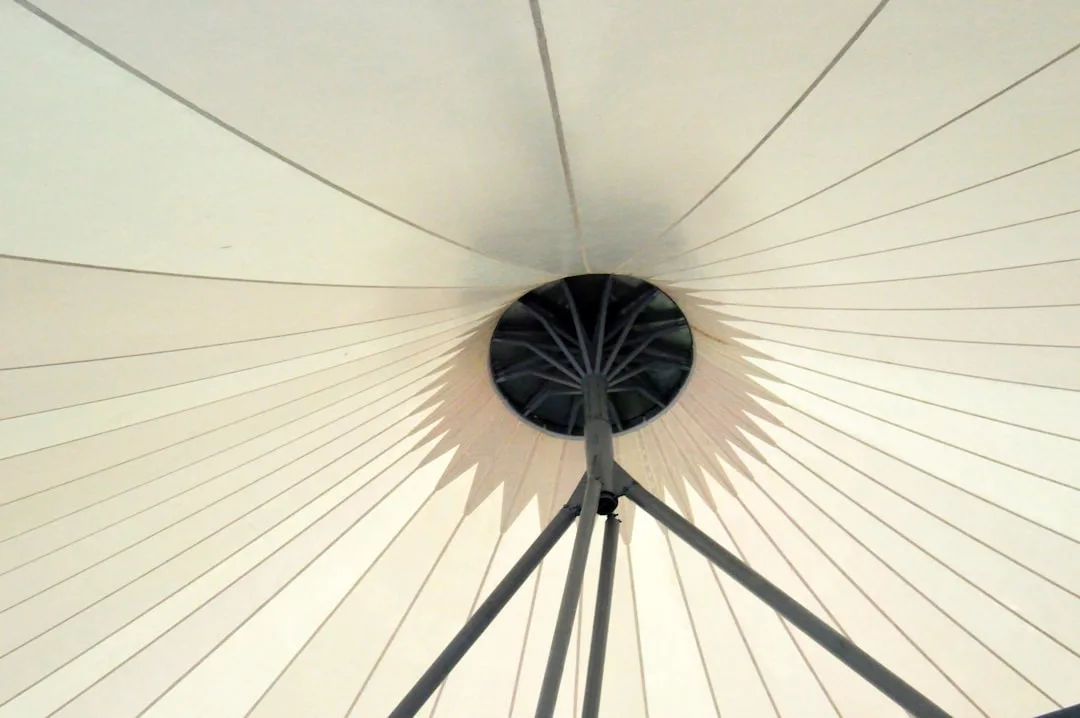 View looking up at the underside of a luxury tensile fabric canopy structure with radial support beams and a central circular hub.