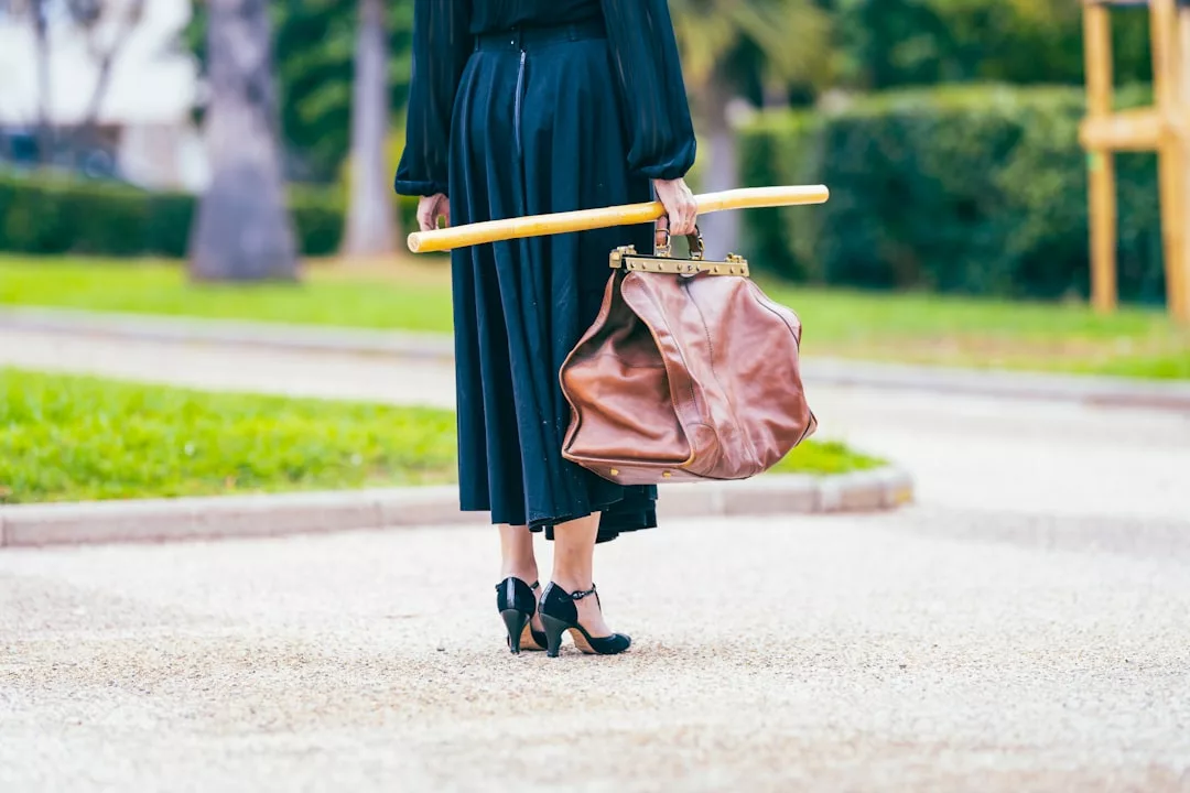 A person in a long black skirt and heels exudes fashion and lifestyle vibes as they stand outdoors, holding a large brown bag and a broomstick over their shoulder.