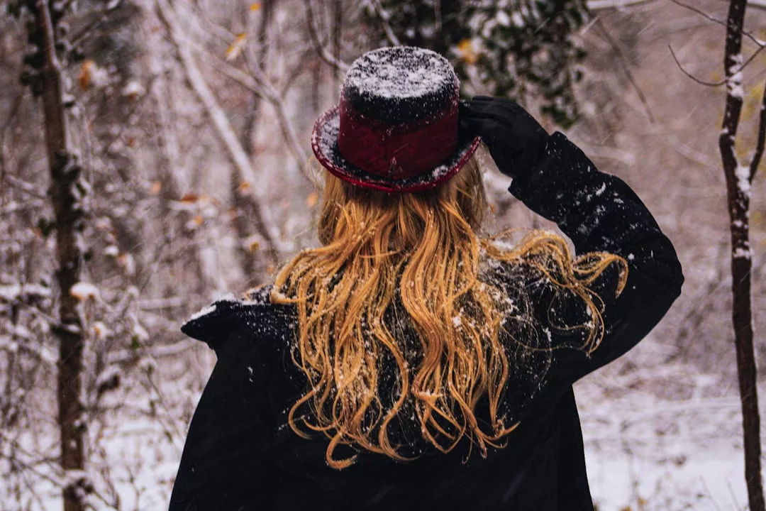 A person with long blonde hair, dressed in a black coat and red hat, stands in a snowy forest. The scene radiates luxury fashion, reminiscent of a Glamesia winter collection viewed from behind.