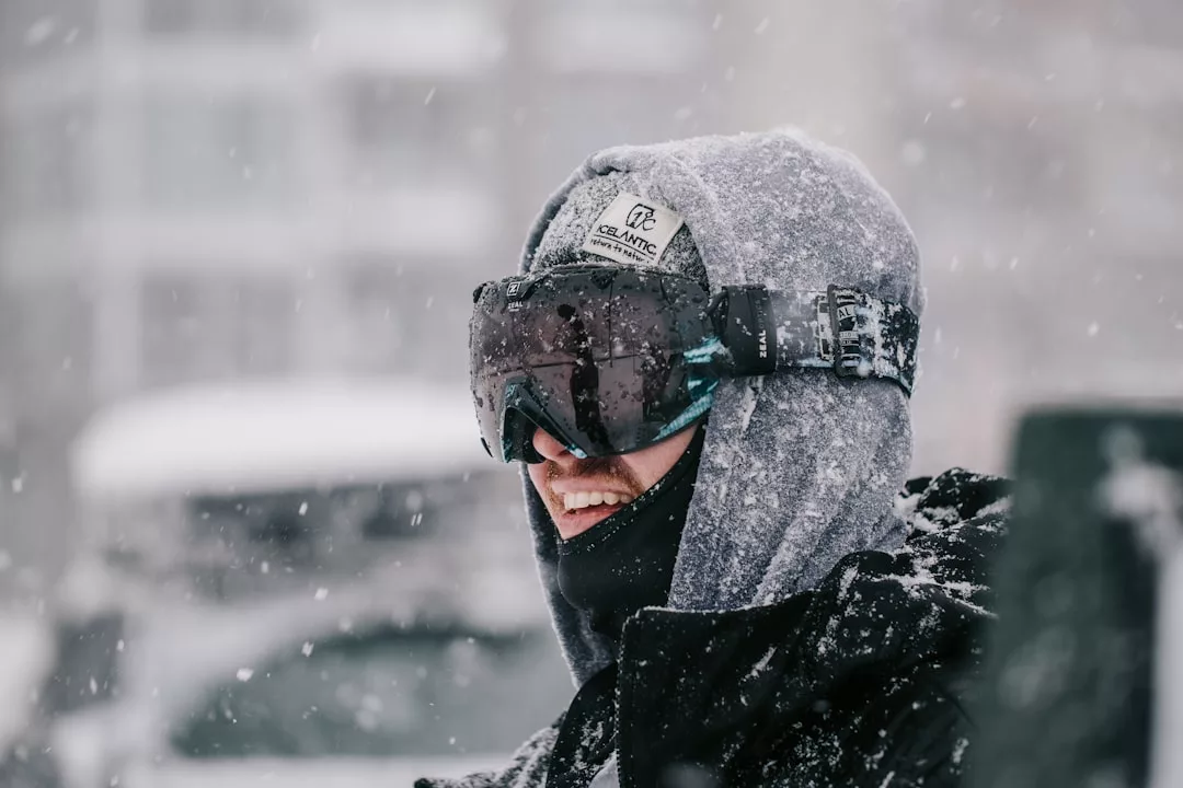 A person wearing a hooded jacket, ski goggles, and a face covering stands outside in falling snow, showcasing Glamesia fashion for the adventurous lifestyle.