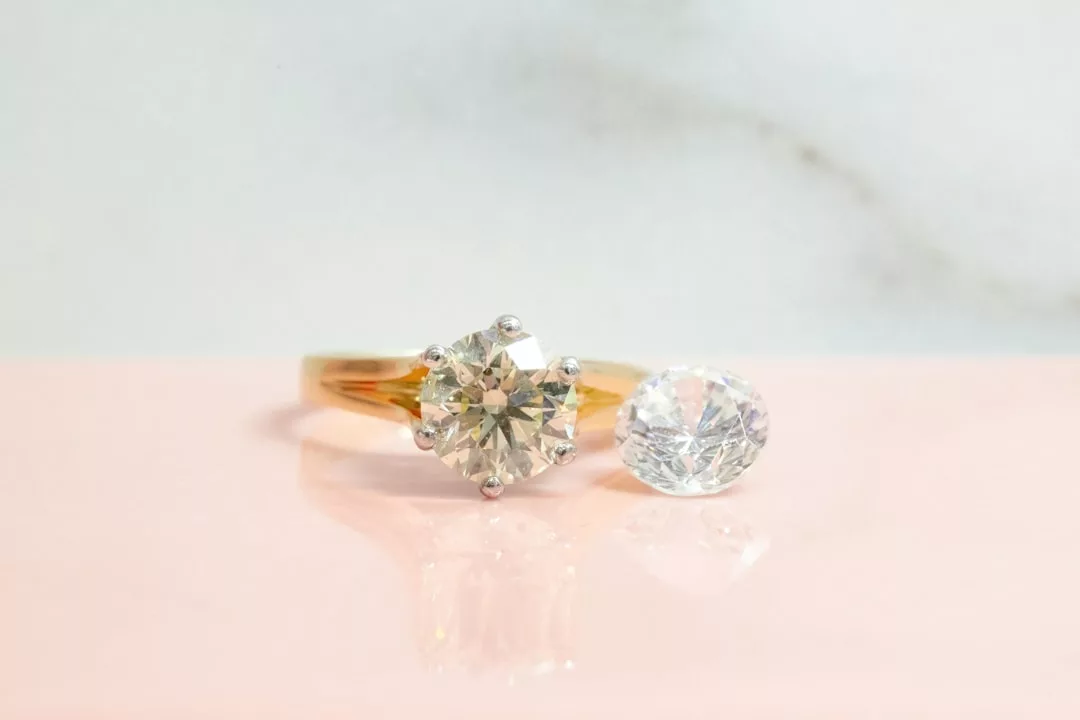 A gold ring with a round solitaire diamond sits next to a loose, similarly cut diamond on a pink and white background, capturing the essence of Glamesia fashion.