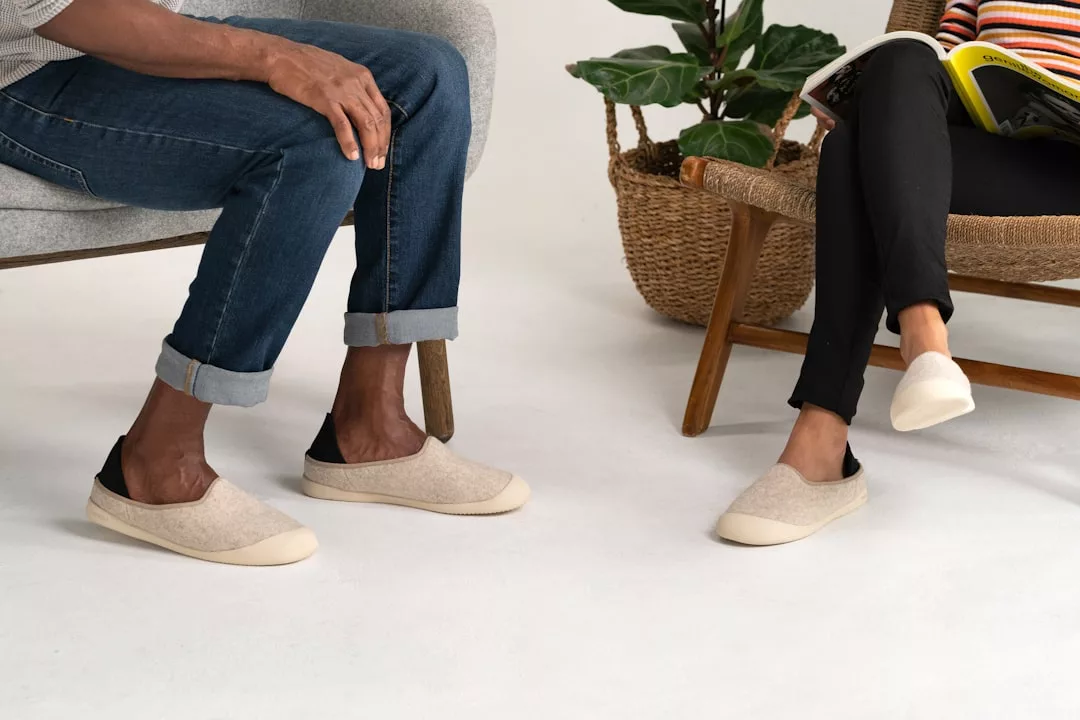 Two people sit on chairs, each wearing light beige Glamesia slip-on shoes. One holds a book or magazine, embracing a relaxed lifestyle; a woven basket with a plant accents the cozy background.