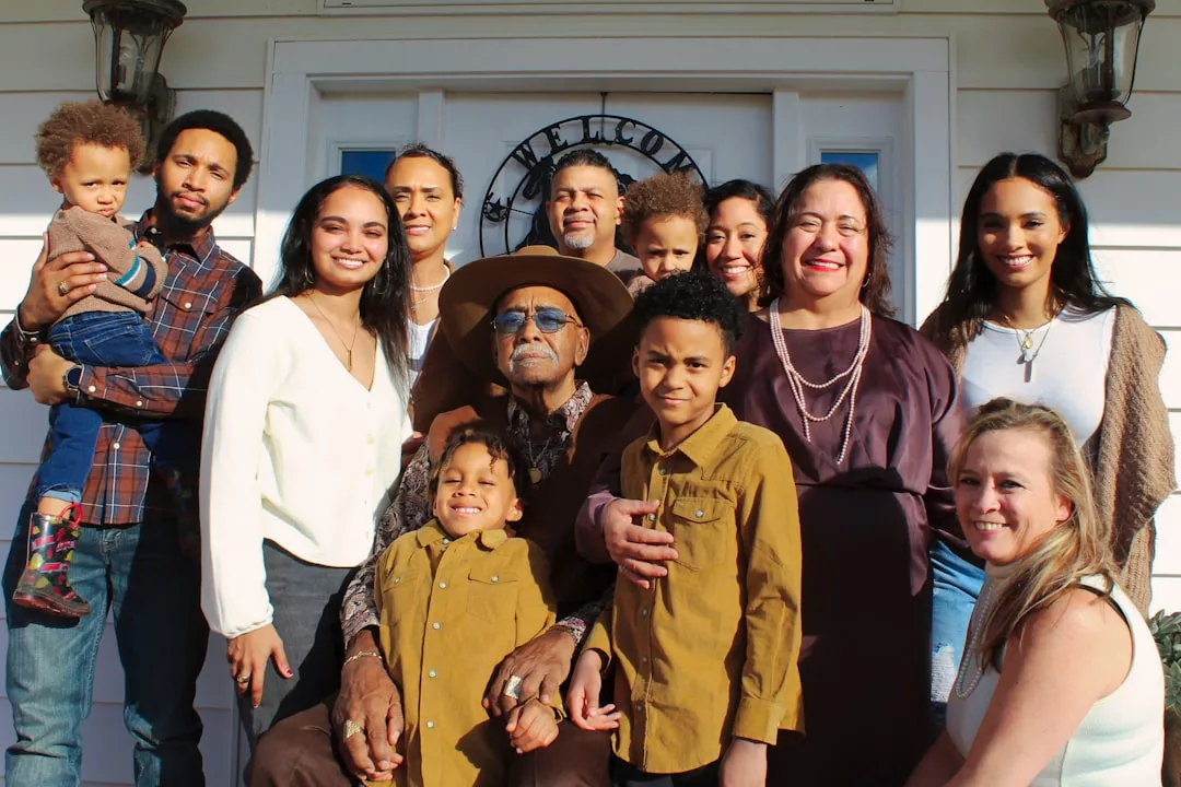 A multi-generational family poses together in front of a luxury house, smiling for the camera on a sunny day.