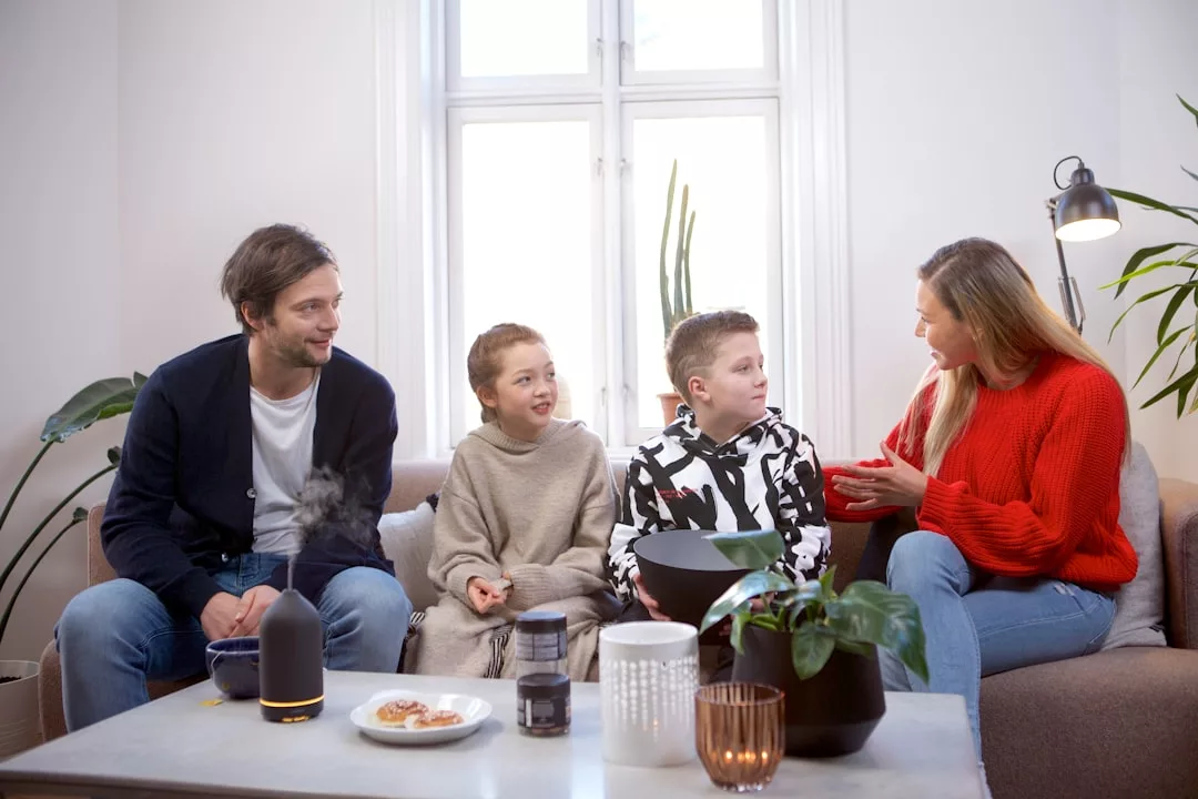 A man, woman, and two children sit together on a sofa in a living room, talking and smiling, enjoying snacks and a plant on the coffee table—a perfect glimpse of relaxed lifestyle and family togetherness.