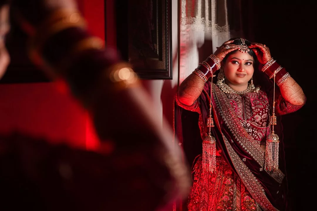 A woman in a Glamesia maroon bridal outfit adjusts her headpiece while standing in front of a mirror, with red lighting illuminating the scene, capturing the essence of fashion and modern laxary.