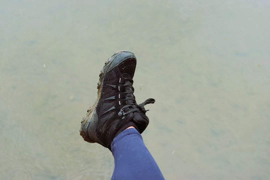 A person wearing blue leggings and a black hiking boot extends their leg over a shallow, murky body of water, capturing a moment where fashion meets an adventurous lifestyle.