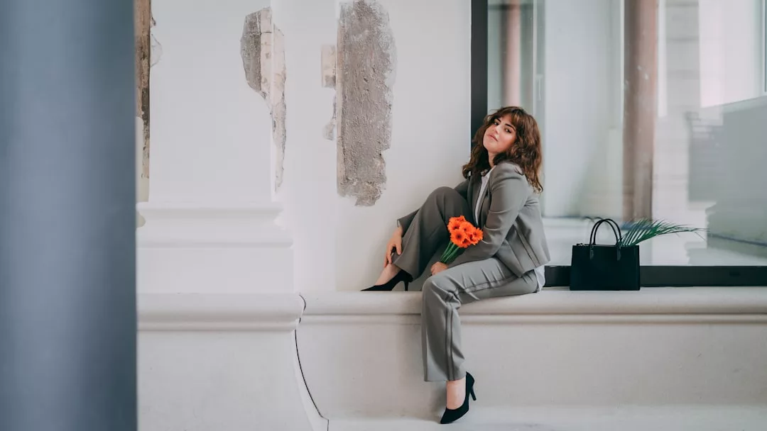 A woman in a gray suit sits on an indoor ledge holding orange flowers, her black handbag and green leaves beside her—a true Glamesia moment in fashion.