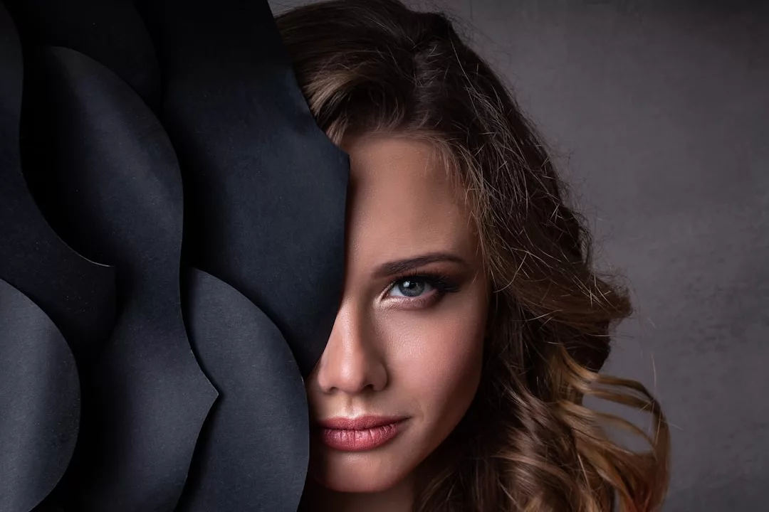A woman with wavy hair gazes at the camera, half of her face veiled by layered black paper shapes, creating a striking Glamesia fashion statement against a neutral background.