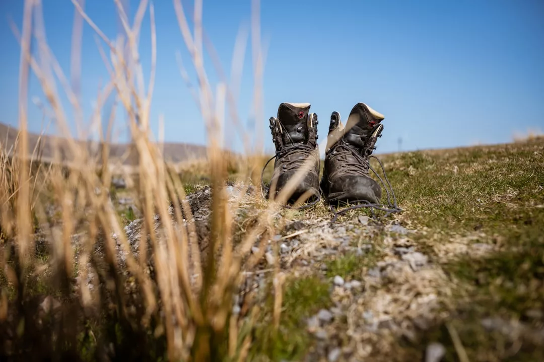 A pair of luxury hiking boots sits upright on grassy ground under a clear blue sky, with dry grass in the foreground.