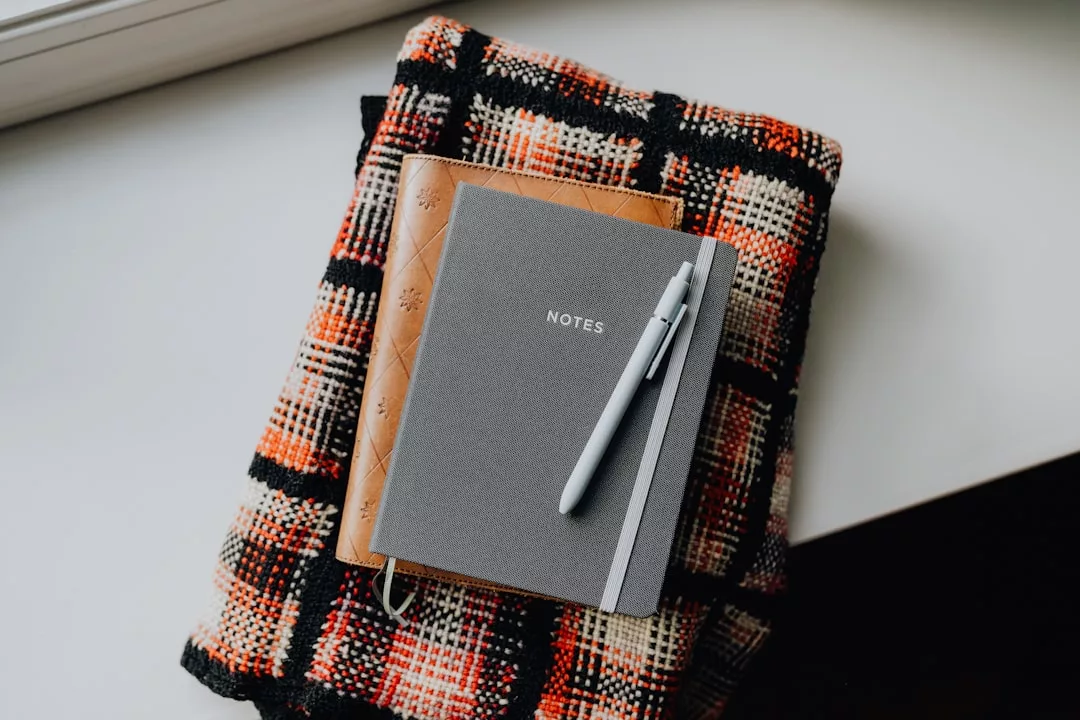 A gray Glamesia notebook with "NOTES" on the cover and a pen rests atop a brown journal and folded plaid fabric, adding a touch of lifestyle luxury to your white surface.
