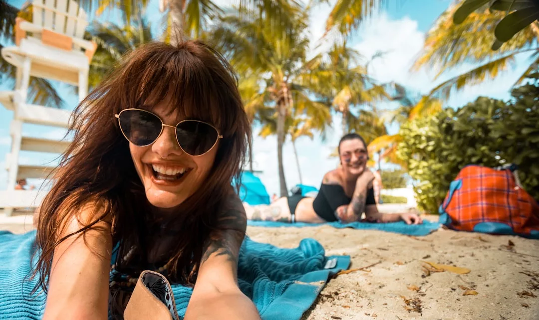 Two people lying on blue towels on a sandy beach, surrounded by palm trees, smiling at the camera under sunny skies, enjoying a touch of Glamesia luxury.